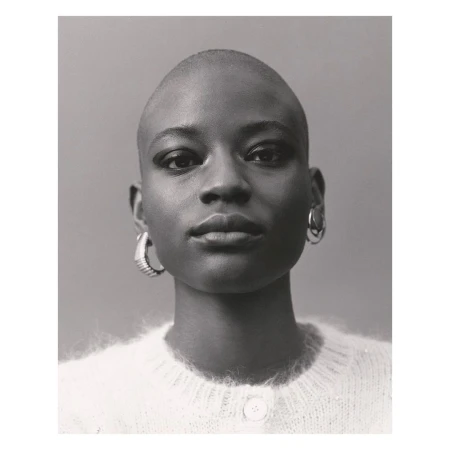 Portrait of a young Black woman with a shaved head, wearing a white sweater and hoop earrings, posing in front of a neutral background.