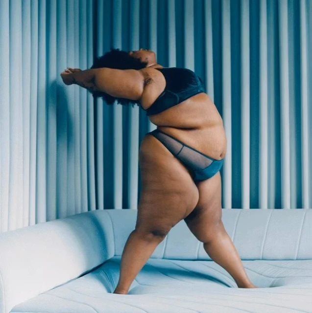 An adult female poses in a teal lingerie set against a blue vertically striped background in a studio setting, lit with studio lighting.