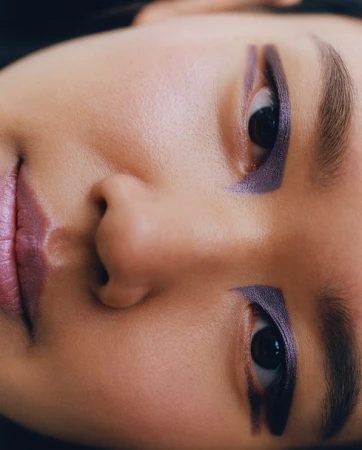 Close-up portrait of a woman's face featuring geometric purple eye makeup and matching mauve lips, photographed by Felicity Ingram.