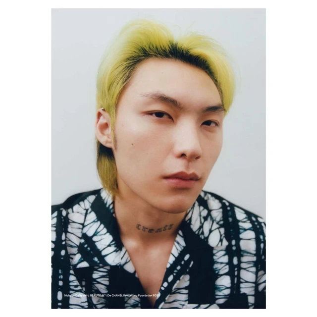 Portrait of an adult male with bright yellow hair, wearing a black and white patterned shirt against a plain light grey background.