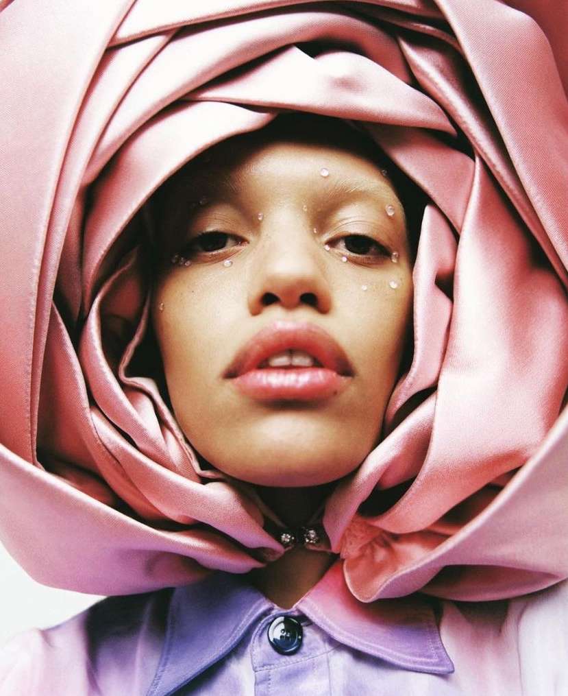 Close-up of an adult female model wearing a rose-shaped head covering with water droplets on her face in a beauty or fashion setting.