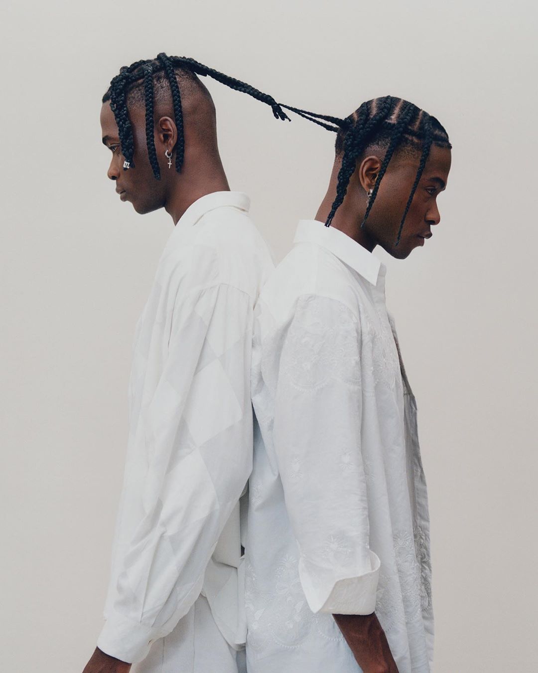 Two Black men stand back to back in a bright, high-key portrait, wearing white button-down shirts and braided hairstyles against a neutral backdrop.