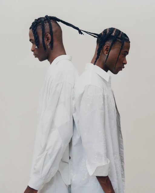 Two Black men stand back to back in a bright, high-key portrait, wearing white button-down shirts and braided hairstyles against a neutral backdrop.