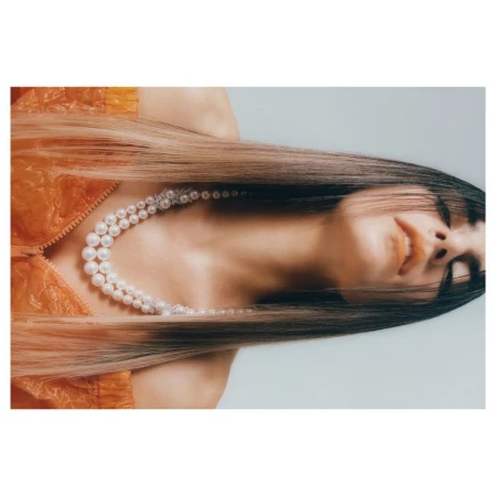 Close-up portrait of a woman wearing an orange puffer jacket and a double-strand pearl necklace against a plain white background.