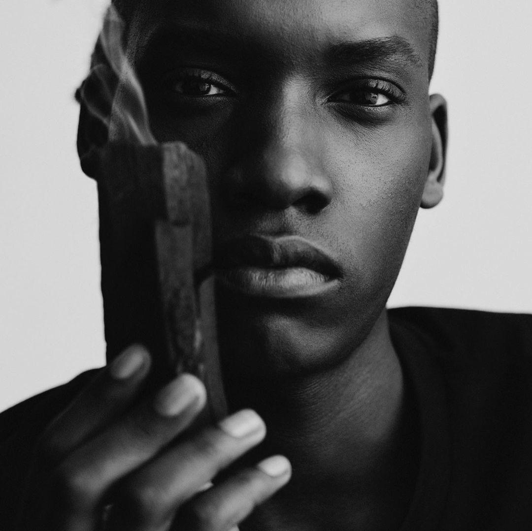 Close-up black and white portrait of a dark-skinned adult man holding smoking incense against a neutral background, emphasizing dramatic lighting.
