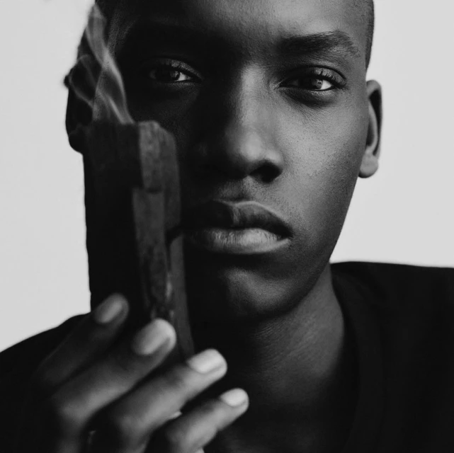 Close-up black and white portrait of a dark-skinned adult man holding smoking incense against a neutral background, emphasizing dramatic lighting.