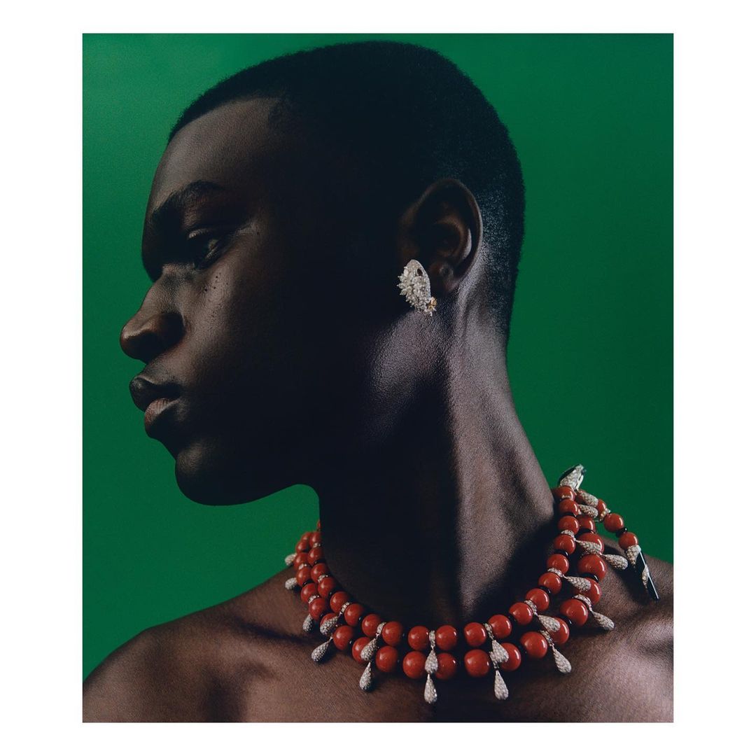 Profile portrait of a Black man wearing a red beaded necklace and a sparkling earring against a vibrant green backdrop.