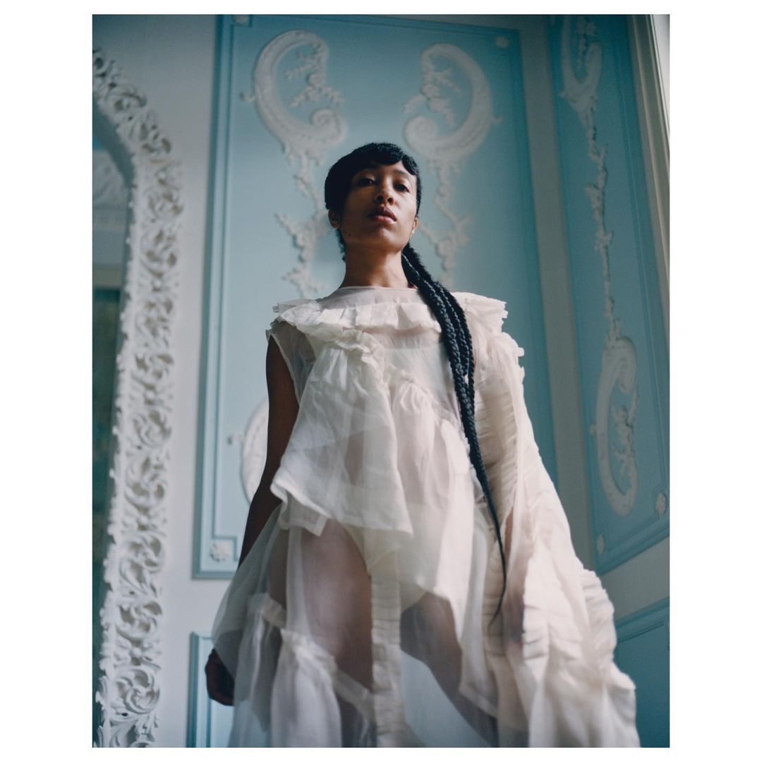 Young woman in a ruffled white dress posing in a room with light blue walls and white ornate trim, portrait orientation.