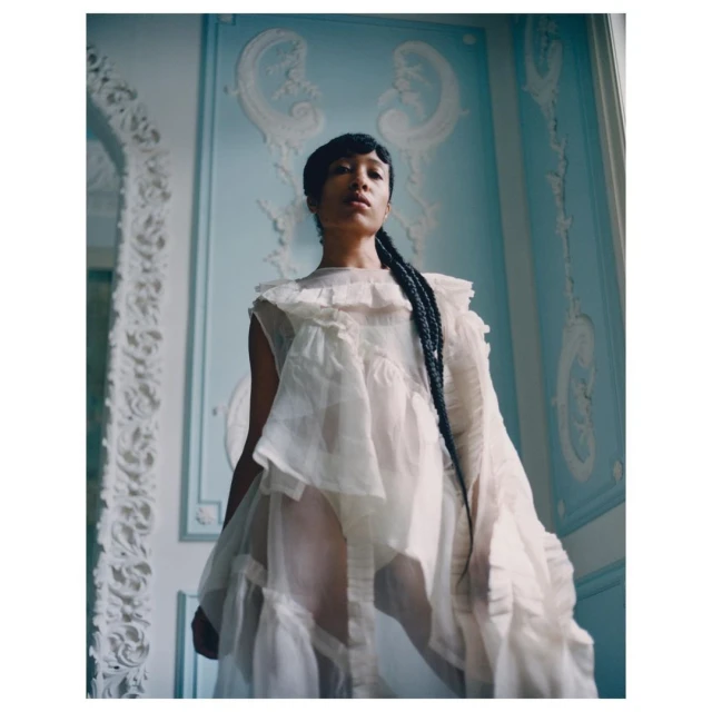 Young woman in a ruffled white dress posing in a room with light blue walls and white ornate trim, portrait orientation.