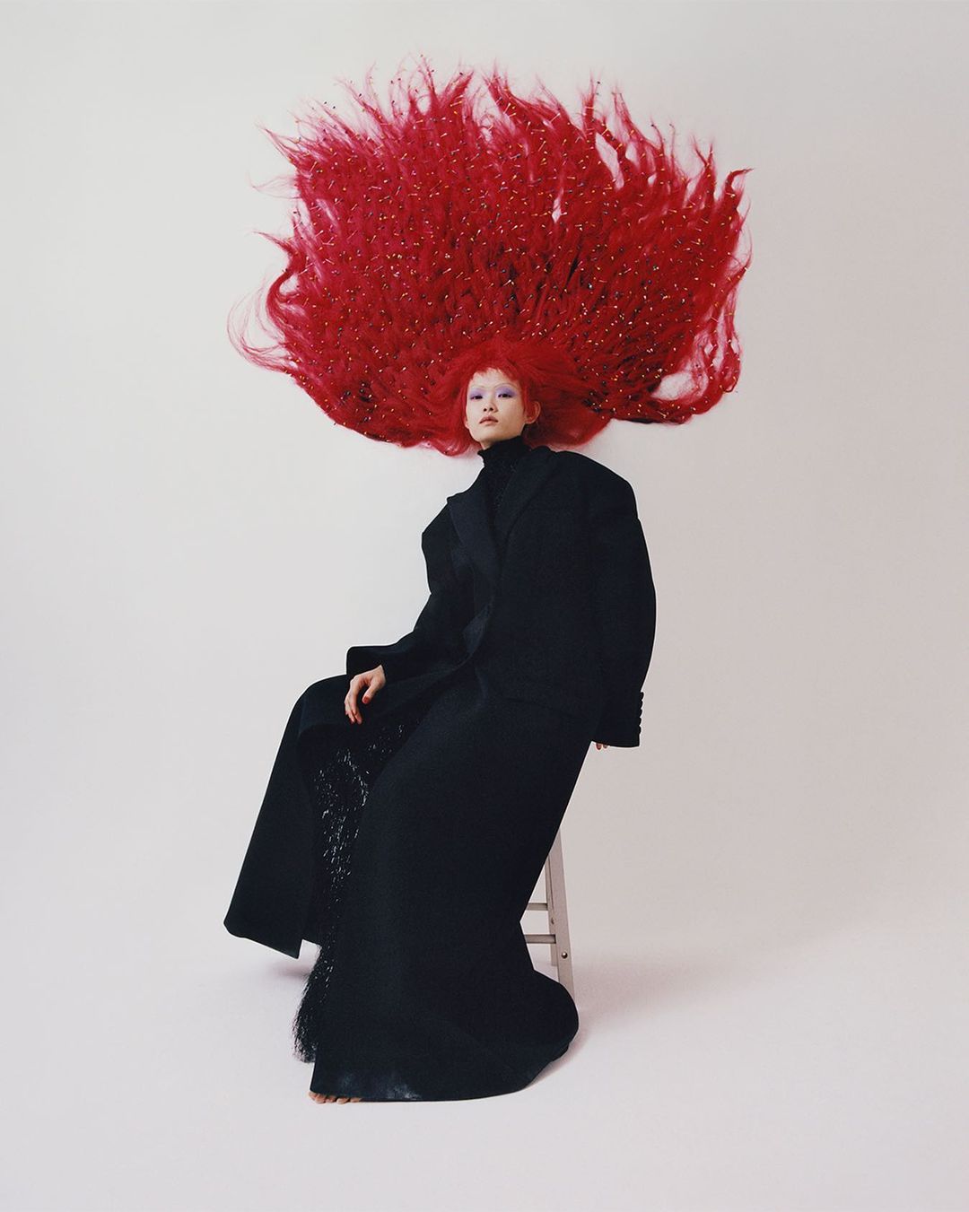 A fair-skinned woman sits on a stool wearing a black gown and sporting voluminous, shiny, red hair, set against a neutral background.