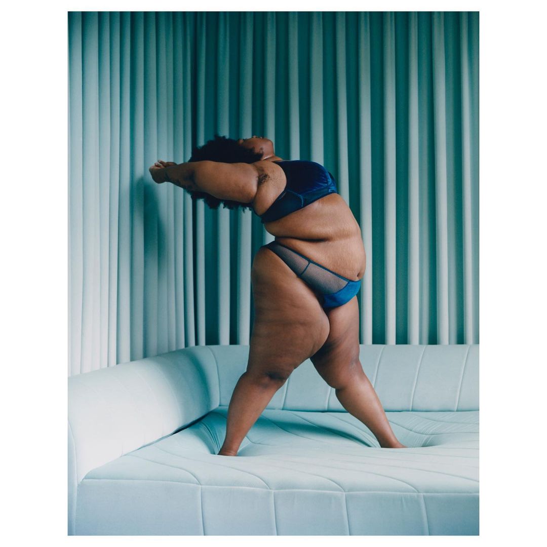 A full-figure female model in lingerie poses against a blue background, arching her back on a white couch in a studio setting.