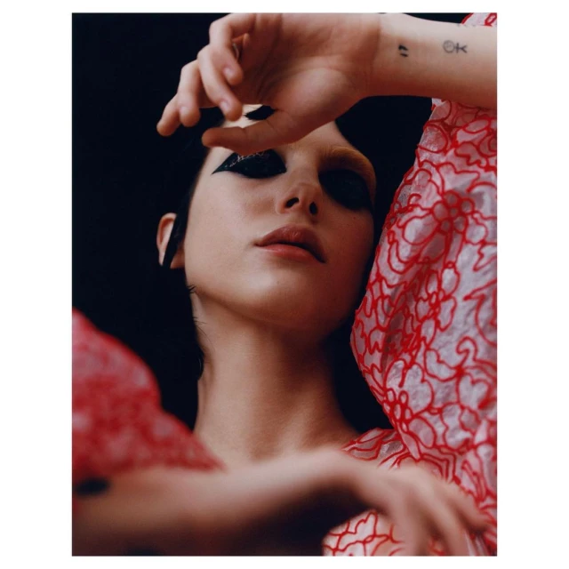 Close-up portrait of a young adult female with dramatic black eye makeup, wearing a red patterned sheer blouse, against a dark backdrop.