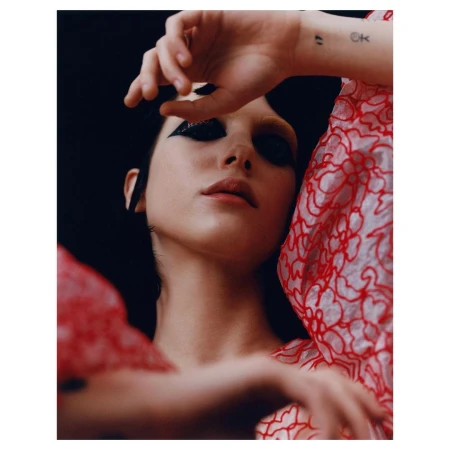 Close-up portrait of a young adult female with dramatic black eye makeup, wearing a red patterned sheer blouse, against a dark backdrop.