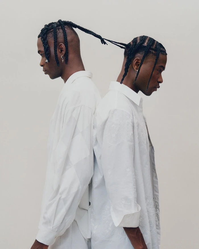 Two black men standing back to back in white shirts with their braided hair tied together against a seamless background