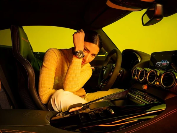 Woman in a yellow sweater and white pants sitting in a black sports car, interior shot, with golden yellow background lighting.
