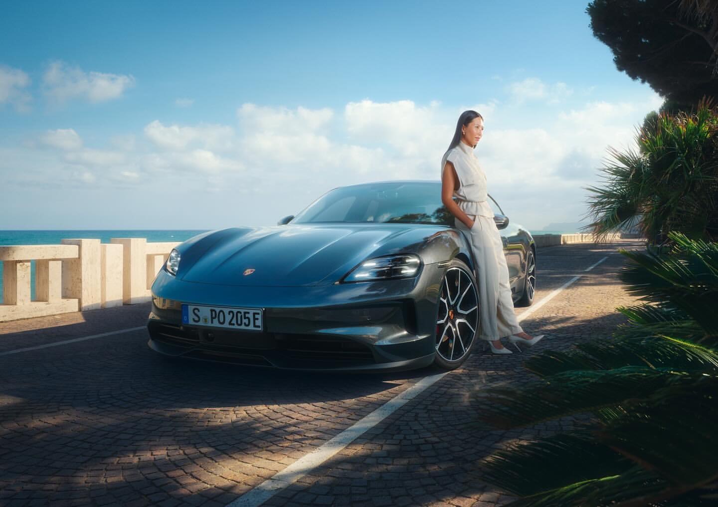 Teal Porsche Taycan electric car parked on a seaside road with an adult woman leaning on it under a sunny sky.