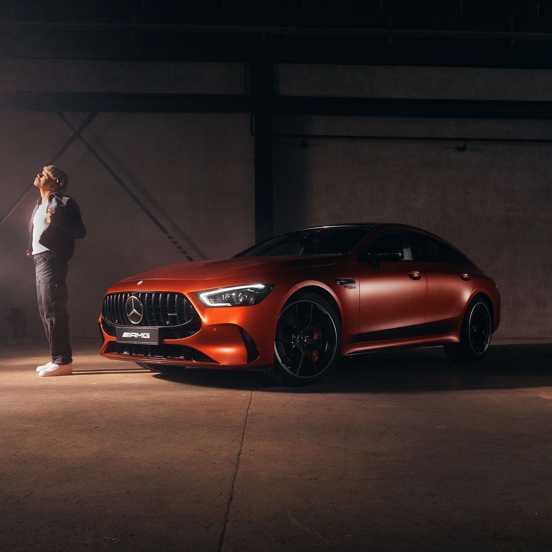 A bronze-colored Mercedes-AMG GT four-door coupe is shown with a man in casual attire standing next to it in a garage setting.