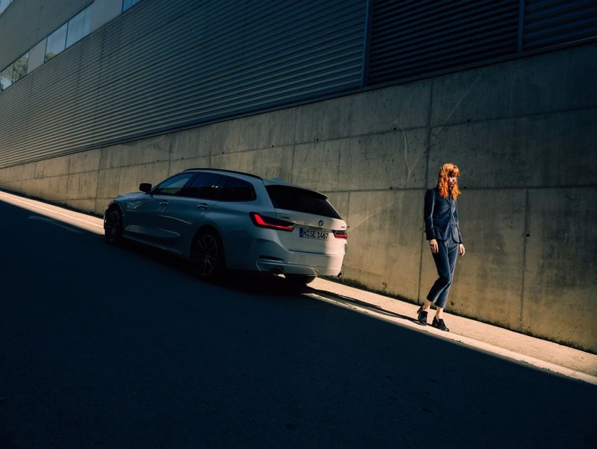 Silver BMW station wagon parked next to an adult female in a blue suit in an urban environment during daylight, with sunlight casting shadows.
