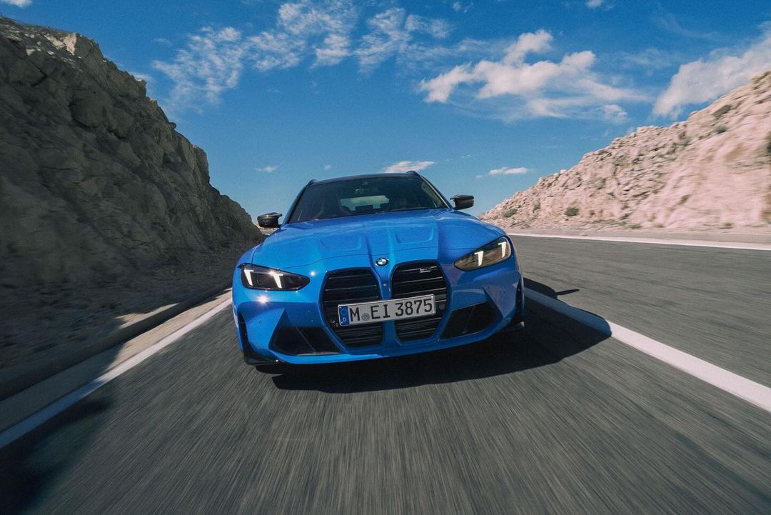 A bright blue BMW M3 Touring drives on an asphalt road framed by mountains under a blue sky with fluffy white clouds.