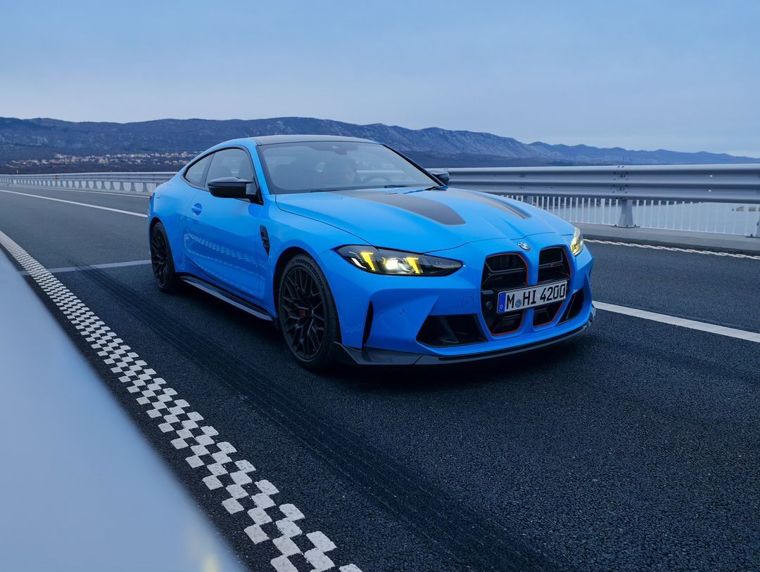 A vibrant blue BMW M4 coupe is parked on a bridge with a mountain range backdrop, showcasing automotive design and scenic beauty.
