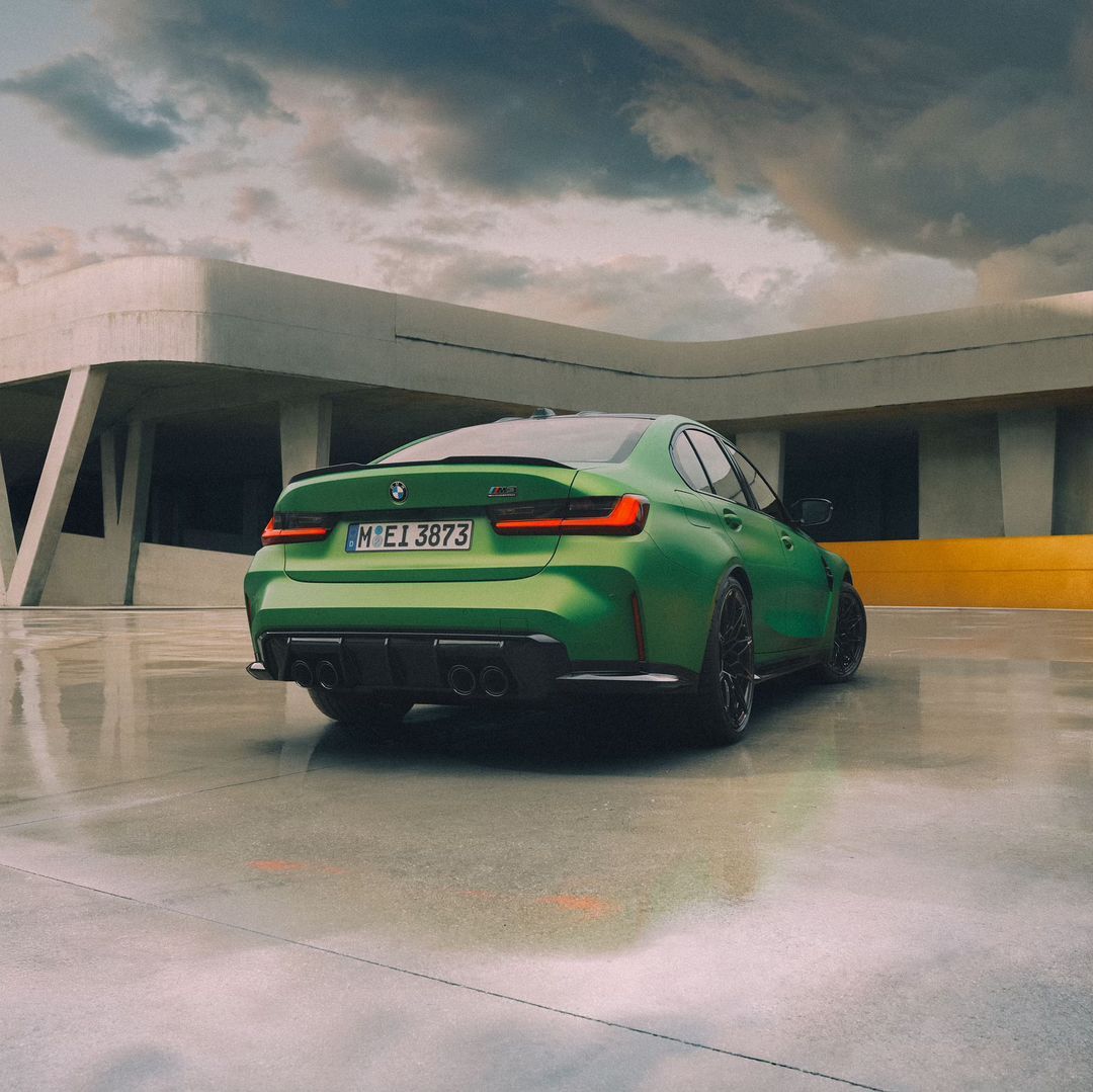 Green BMW M3 automobile with black wheels and spoiler parked outside a modern architectural building under a cloudy sky.