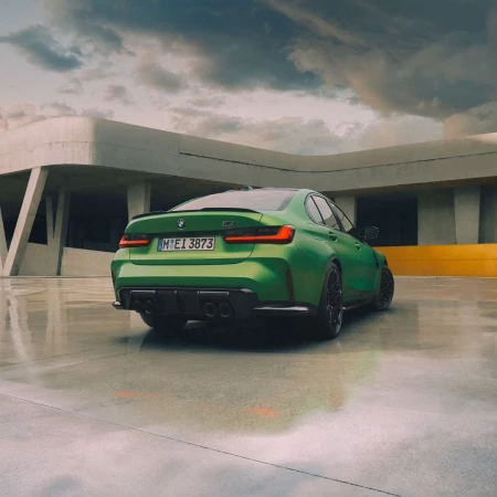 Green BMW M3 automobile with black wheels and spoiler parked outside a modern architectural building under a cloudy sky.