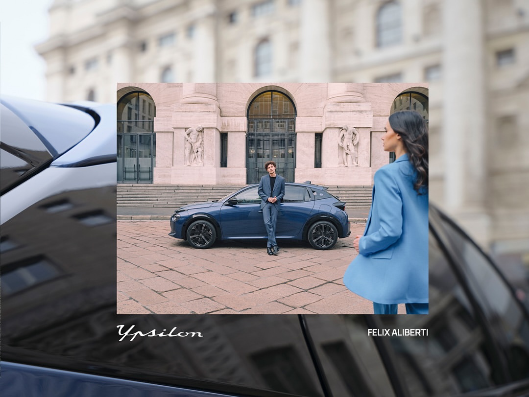Blue car showcased outdoors with two people: a man leaning on the car and a woman in a blue suit, set against a city building.