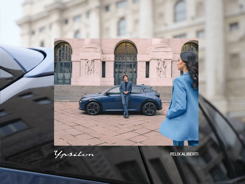 Blue car showcased outdoors with two people: a man leaning on the car and a woman in a blue suit, set against a city building.