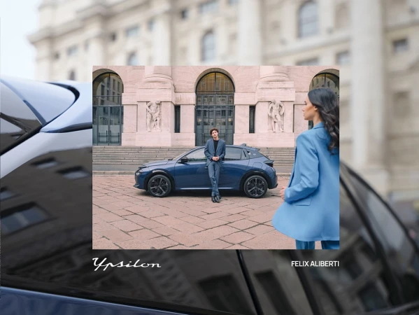 Blue car showcased outdoors with two people: a man leaning on the car and a woman in a blue suit, set against a city building.