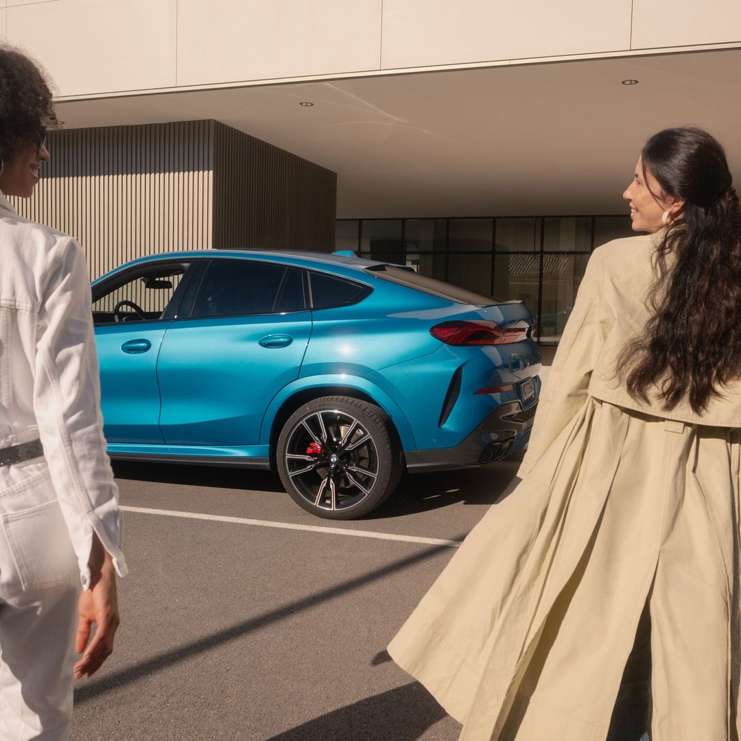 A blue BMW X6 coupe SUV is parked outdoors, with two adult women walking on either side of it on a sunny day.