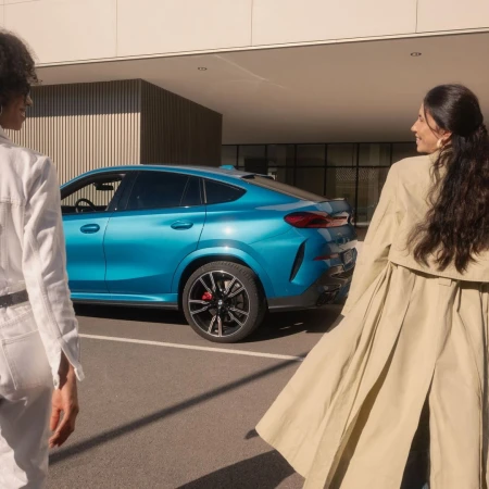 A blue BMW X6 coupe SUV is parked outdoors, with two adult women walking on either side of it on a sunny day.