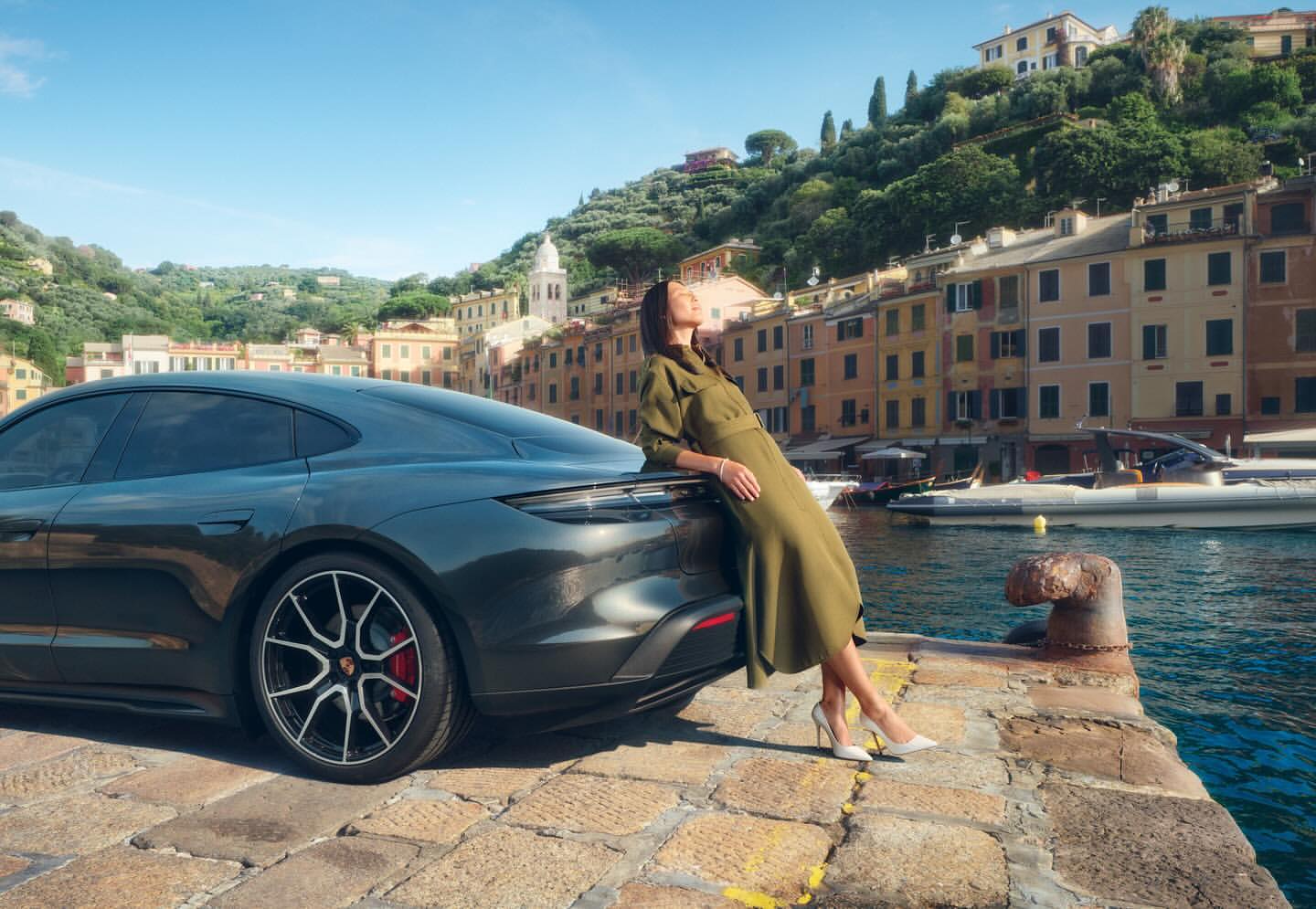 A dark grey Porsche Taycan parked on a cobblestone pier with a woman in a green dress leaning on it, set against a scenic coastal town.