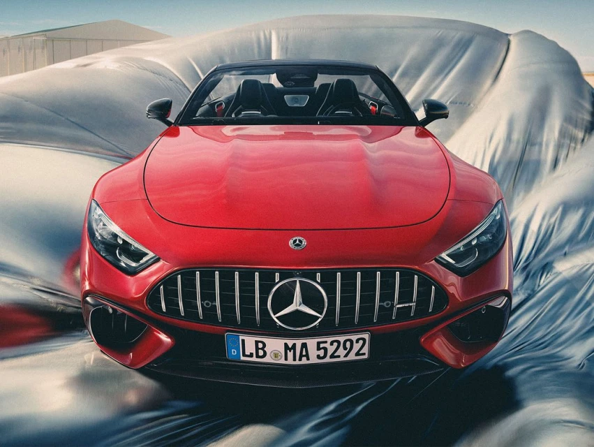 Front-facing view of a red Mercedes-AMG SL roadster with a soft convertible top, partially covered by a gray textile, outdoors.