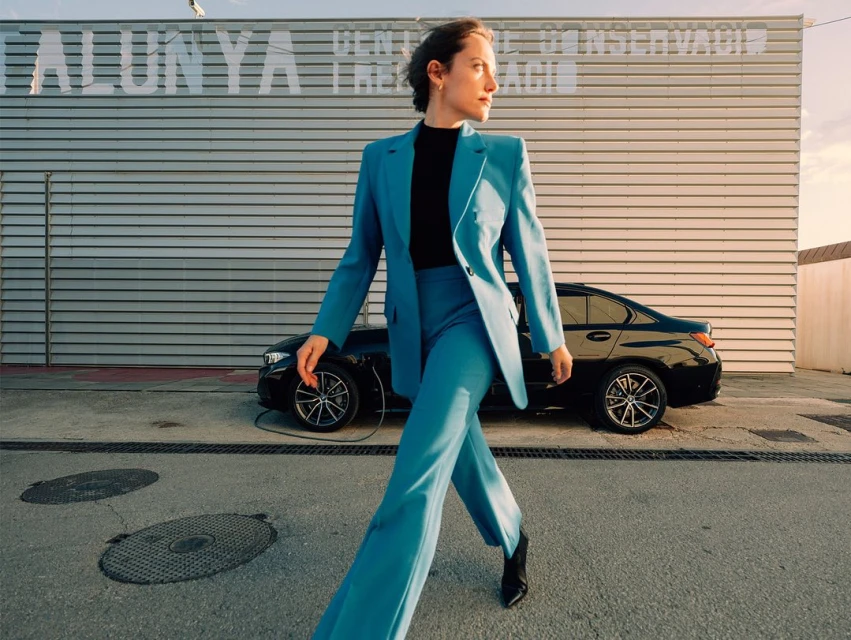 Woman in a turquoise suit walks past a black electric car and an industrial building, photographed in outdoor daylight.
