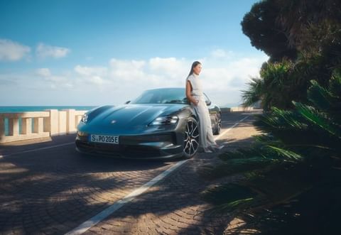 Elegant woman in white leaning on a gray Porsche Taycan electric car near a coastal road with the sea and sky in the background.