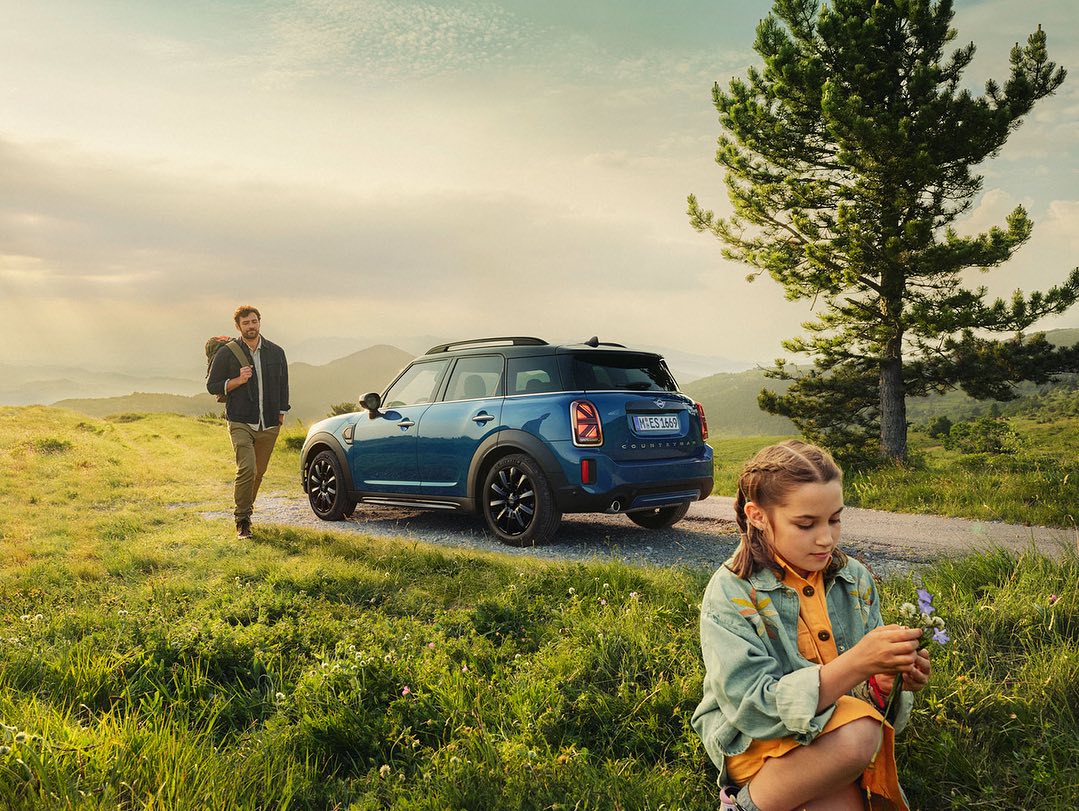 A young girl squats examining a purple flower, a blue Mini Cooper and a man with a backpack behind her on a grassy, mountain road.