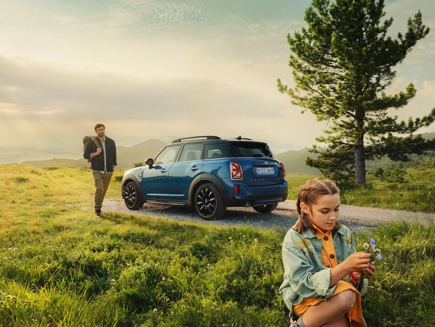 A young girl squats examining a purple flower, a blue Mini Cooper and a man with a backpack behind her on a grassy, mountain road.