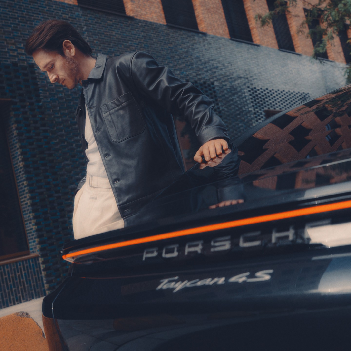 Stylish adult male in a black jacket stands next to a dark Porsche Taycan 4S car, with a modern building visible in the background.