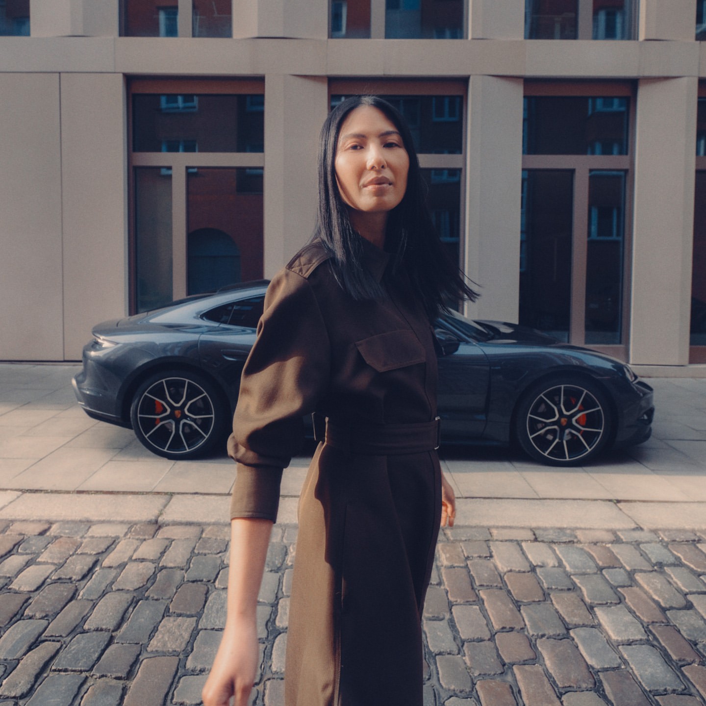 Woman poses in front of a grey Porsche near a building with brick road. The sleek car mirrors her style and the urban setting.