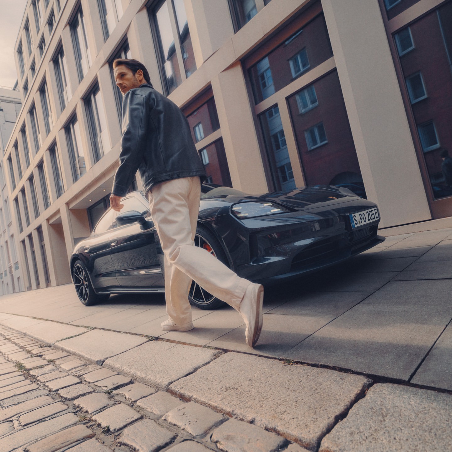 A fashionable adult male walks on a cobblestone street in an urban area, with a dark sports car parked in front of a modern building.