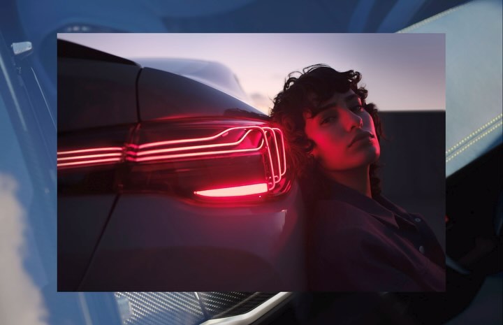 Portrait of a young adult leaning against a car, illuminated by its red taillights, set against a dusk sky. Automotive visual.