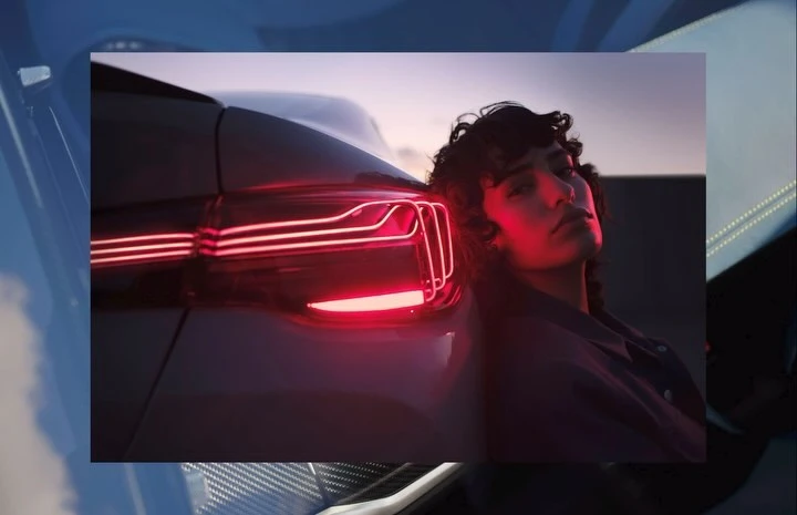 Portrait of a young adult leaning against a car, illuminated by its red taillights, set against a dusk sky. Automotive visual.