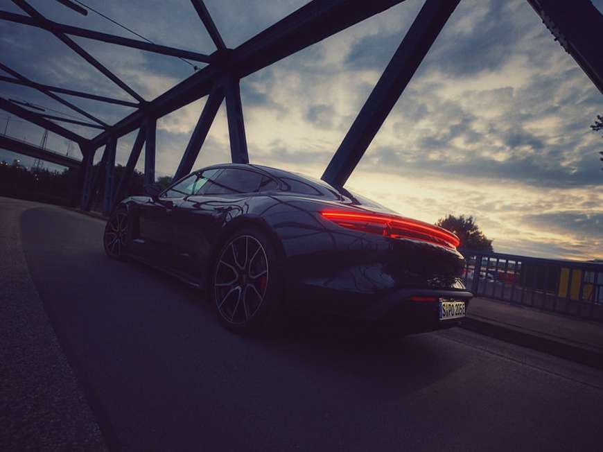 Sleek black Porsche Taycan parked on a bridge against a cloudy sky, showcasing its taillight design and elegant silhouette.