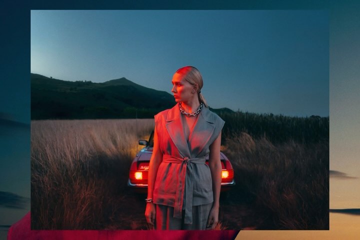 An adult woman stands in front of a vintage car in a field with mountains in the background, captured in a fashion editorial style.