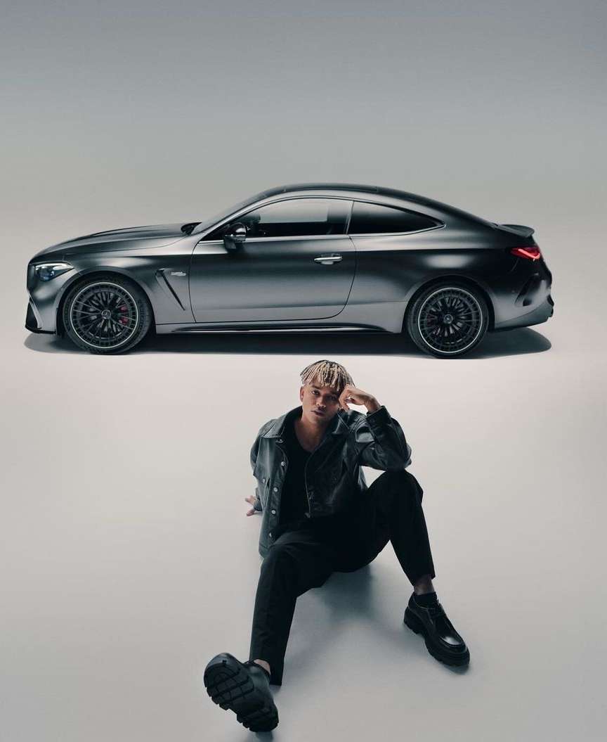 Grey AMG coupe parked indoors with a young adult sitting in front wearing a dark jacket and pants in a studio lighting setup.