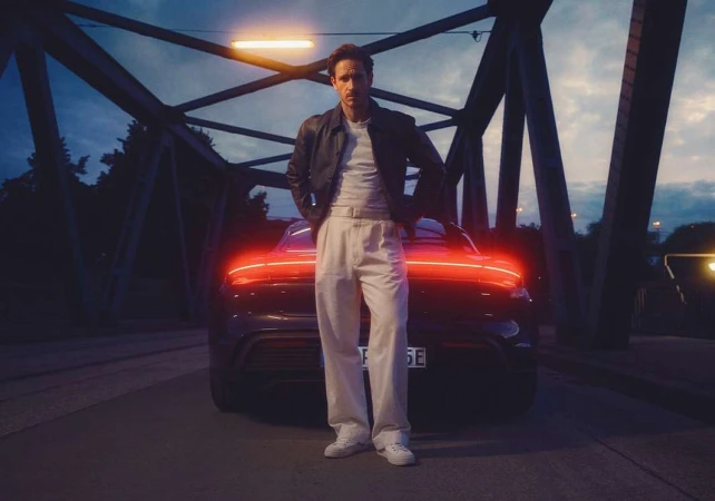 Man in leather jacket posing with black electric car at night beneath a bridge. Automotive fashion editorial shot.