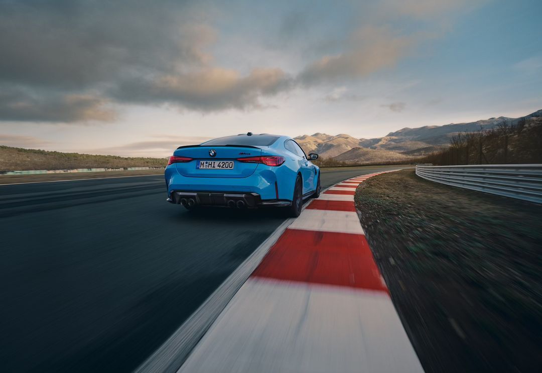 Light-blue BMW M4 coupe driving on a race track with red and white curbing, mountain range and cloudy sky in the background.