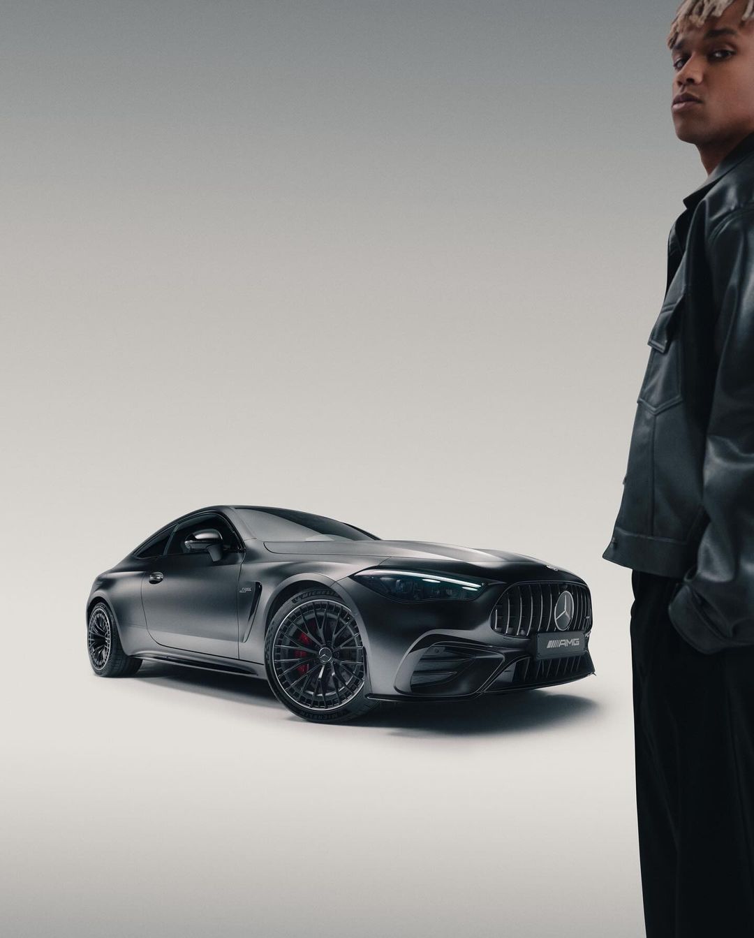 A black Mercedes-AMG GT sports car is showcased alongside a male model with dyed blonde hair in a bright, neutral studio setting.