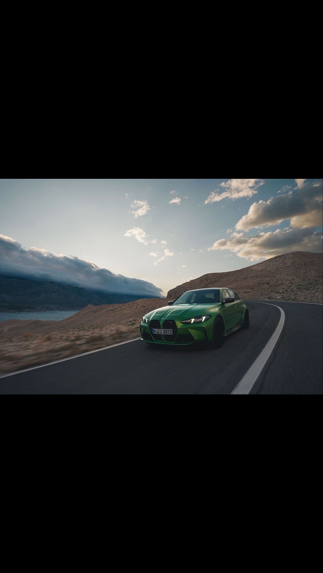 A green BMW car drives on a winding asphalt road with mountains and a lake in the background under a blue and cloudy sky at daytime.