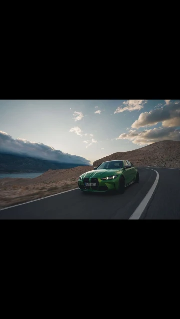 A green BMW car drives on a winding asphalt road with mountains and a lake in the background under a blue and cloudy sky at daytime.